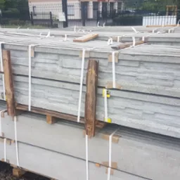 Paleta z prefabrykowanymi, betonowymi elementami ogrodzeniowymi z dekoracyjnym wzorem kamienia, zabezpieczona drewnianymi przekładkami i białymi taśmami, gotowa do transportu.