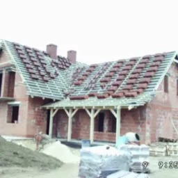 Niedokończony dom z czerwonej cegły z częściowo ułożoną dachówką, widoczne drewniane elementy konstrukcji dachu i podtrzymujące wejście, na pierwszym planie hałda ziemi i materiały budowlane.