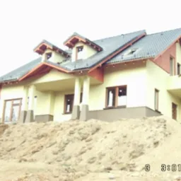 Żółty dom z ciemnoszarą dachówką i drewnianymi elementami wykończeniowymi, częściowo zasłonięty przez usypaną ziemię, z widoczną osobą na balkonie.