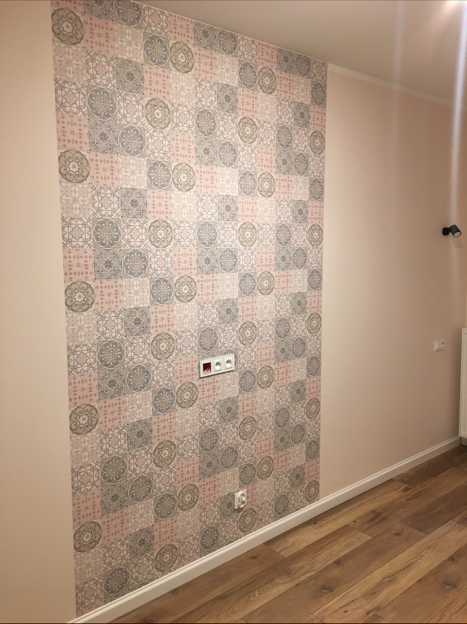 Ściana wyłożona dekoracyjną tapetą w pastelowe wzory, sąsiadująca z gładką, pomalowaną na różowo ścianą, z widocznymi gniazdkami elektrycznymi i drewnianą podłogą.