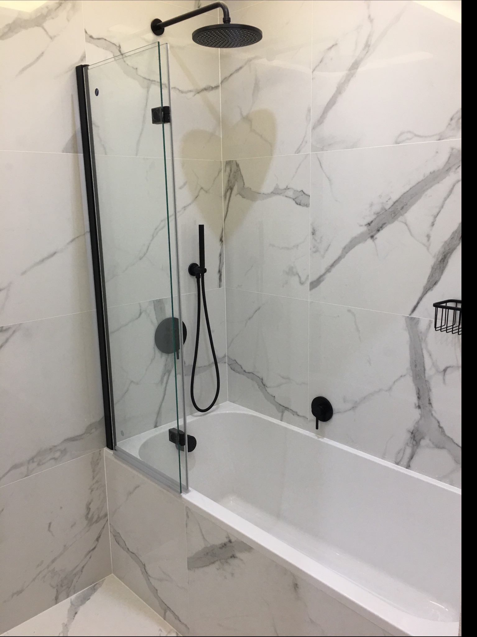 Biała wanna z czarną armaturą i szklanym parawanem na tle płytek imitujących marmur.