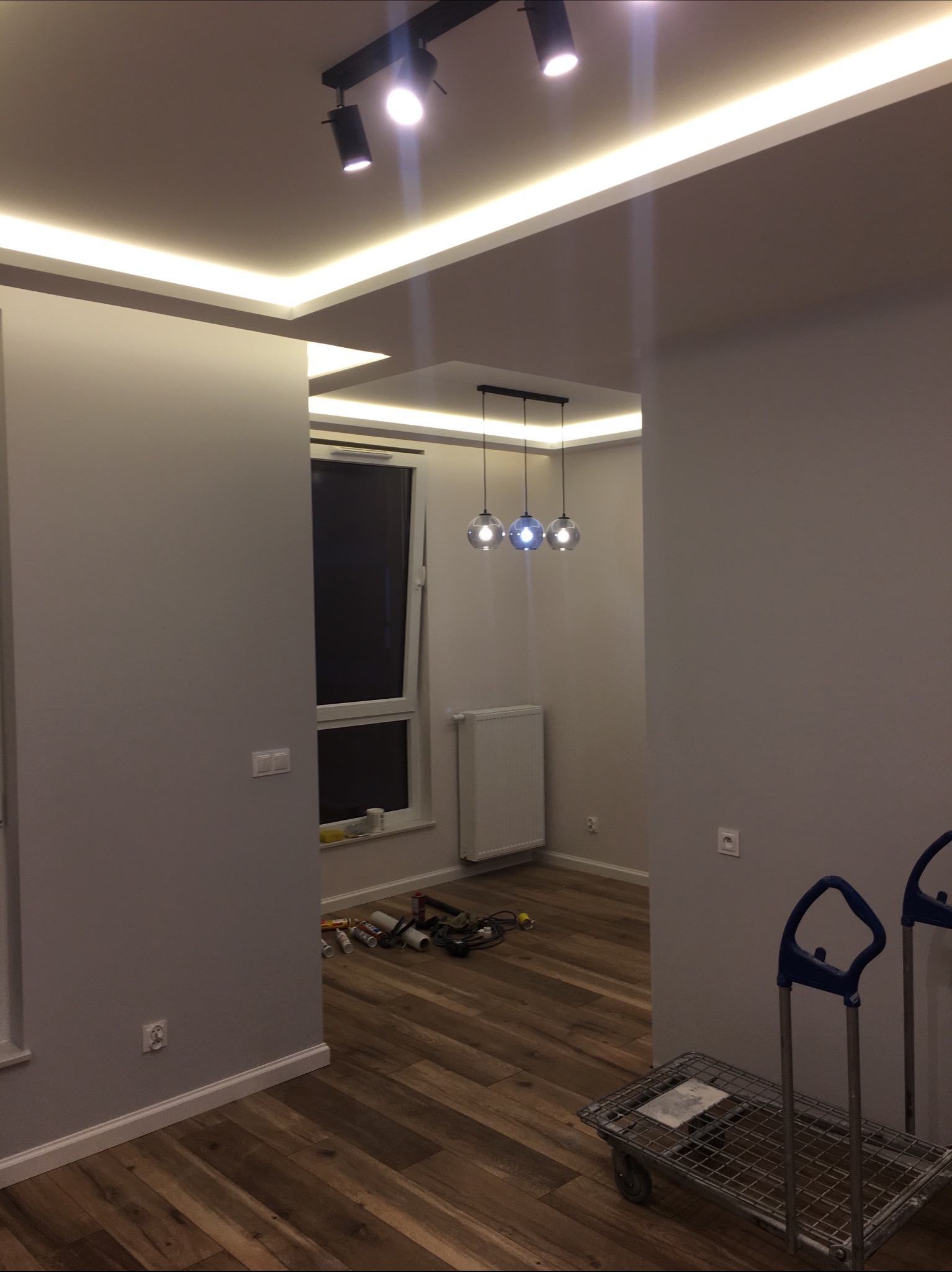 Pokój po odświeżeniu z drewnianą podłogą, szarą ścianą, oświetleniem LED w suficie podwieszanym i wiszącą lampą z trzema szklanymi kloszami, na podłodze widoczne narzędzia i wózek transportowy.