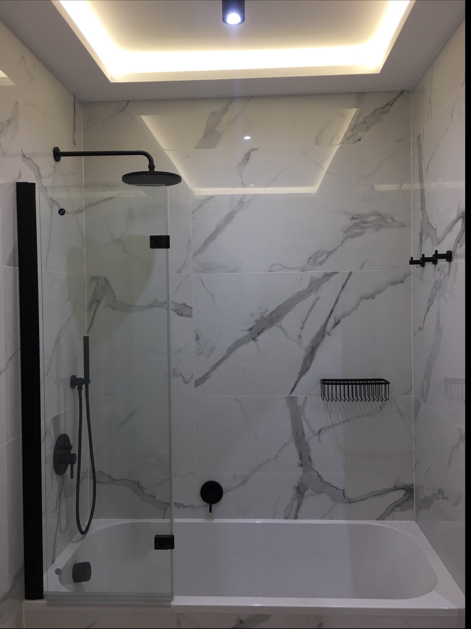 Nowoczesna łazienka z wanną i szklanym parawanem, wykończona białymi płytkami imitującymi marmur i czarną armaturą, oświetlenie sufitowe LED.