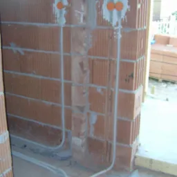 Instalacja elektryczna w ścianie z czerwonej cegły dziurawki, z widocznymi rurkami instalacyjnymi i pomarańczowymi puszkami. Surowy stan, w trakcie prac wykończeniowych.