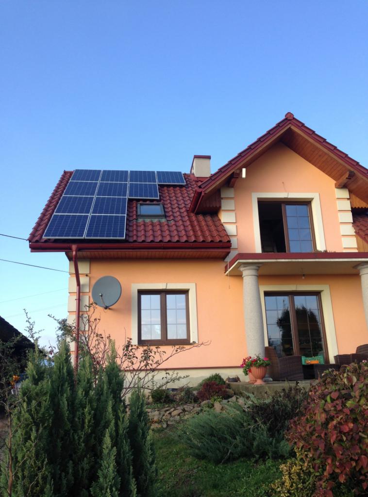 Dom z panelami słonecznymi na czerwonym dachu, antena satelitarna na elewacji. Ujęcie z dołu, błękitne niebo w tle, widoczne krzewy w ogrodzie.