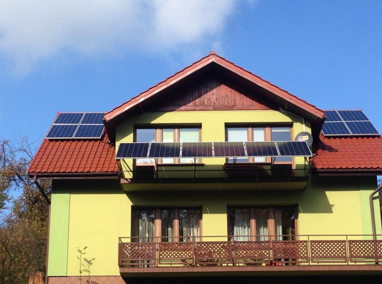 Żółty dom z czerwonym dachem i panelami słonecznymi zamontowanymi na dachu i balkonie, widok z dołu na błękitne niebo w słoneczny dzień.