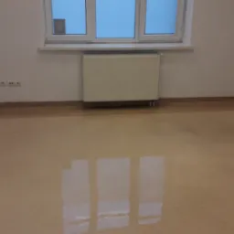 Wypolerowana podłoga z linoleum w pustym biurze, widoczne gniazdka elektryczne i grzejnik pod oknem.