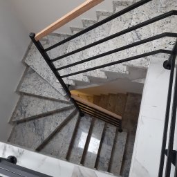 AgAd Bau - Spiralne schody wyłożone jasnym kamieniem z czarną metalową balustradą i drewnianym poręczą, widok z góry.