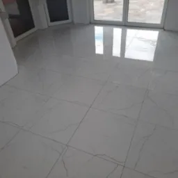 Błyszczące, białe płytki podłogowe imitujące marmur z szarymi żyłkami ułożone w regularną siatkę, odbijające światło z okna balkonowego.