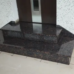 Polerowane, brązowe, dwustopniowe schody wewnętrzne z kamienia, prowadzące do drzwi w Przystajni. Widoczna struktura kamienia i odbicie światła na powierzchni.