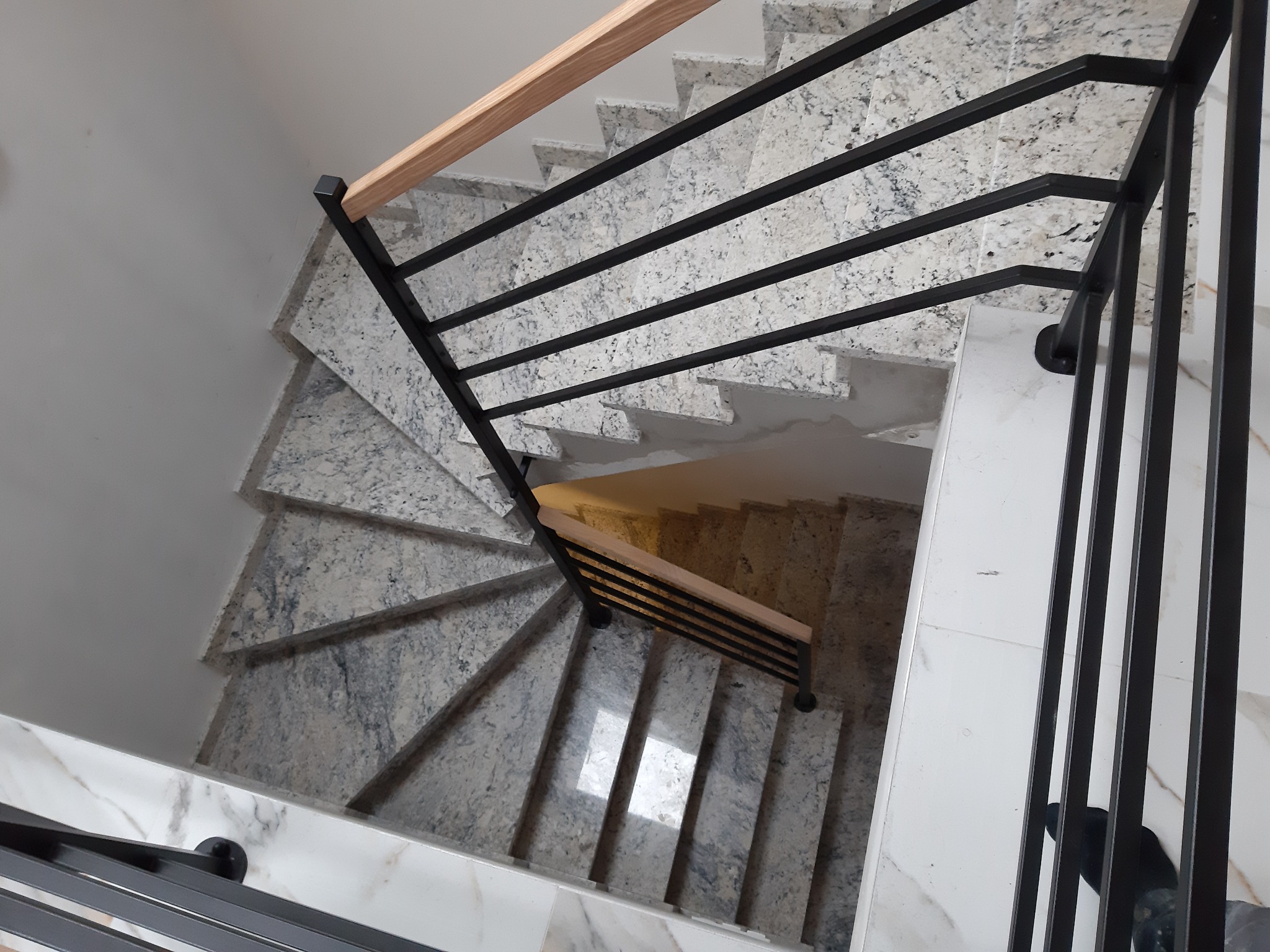 Spiralne schody wyłożone jasnym kamieniem z czarną metalową balustradą i drewnianym poręczą, widok z góry.