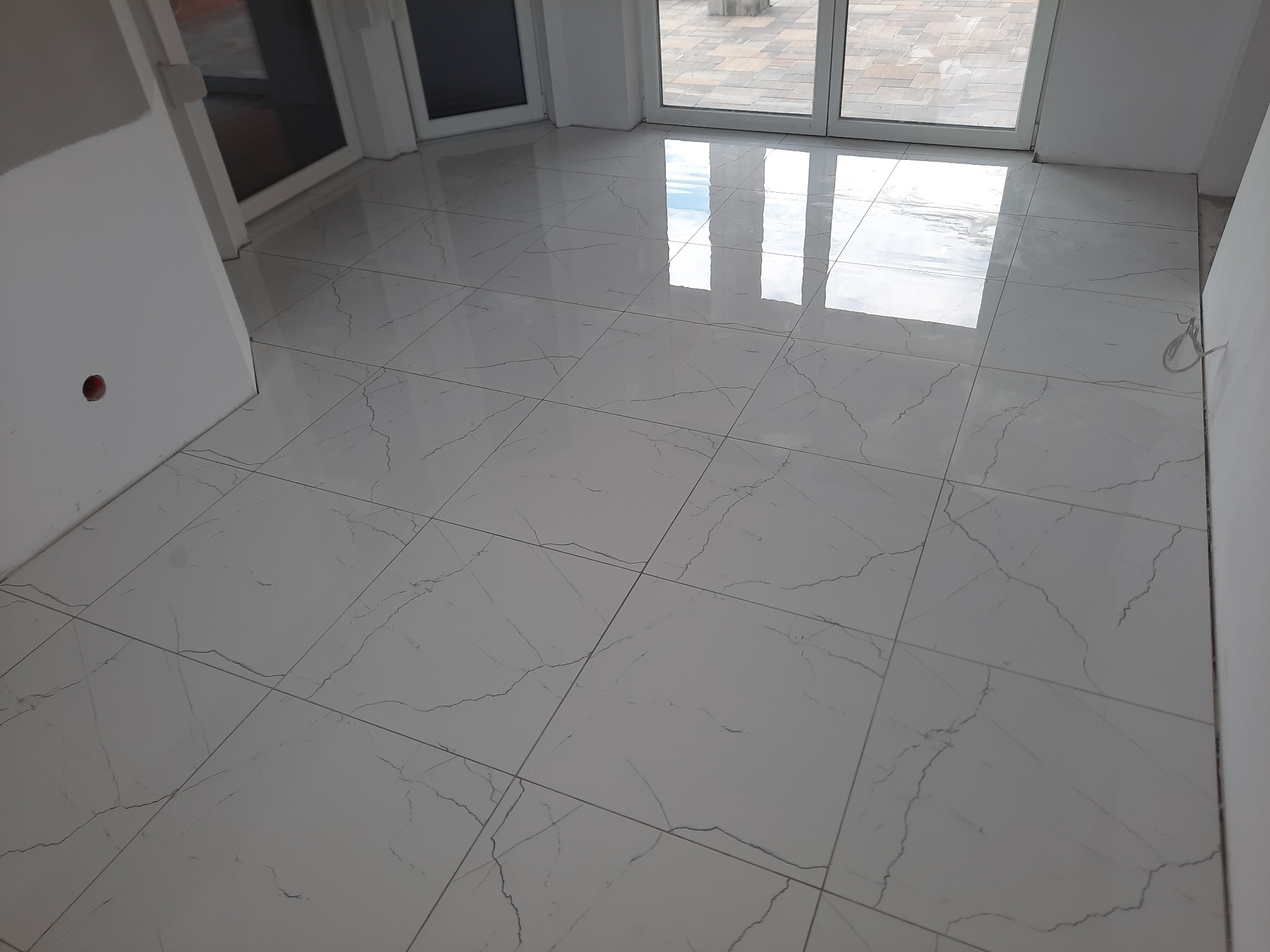 Błyszczące, białe płytki podłogowe imitujące marmur z szarymi żyłkami ułożone w regularną siatkę, odbijające światło z okna balkonowego.