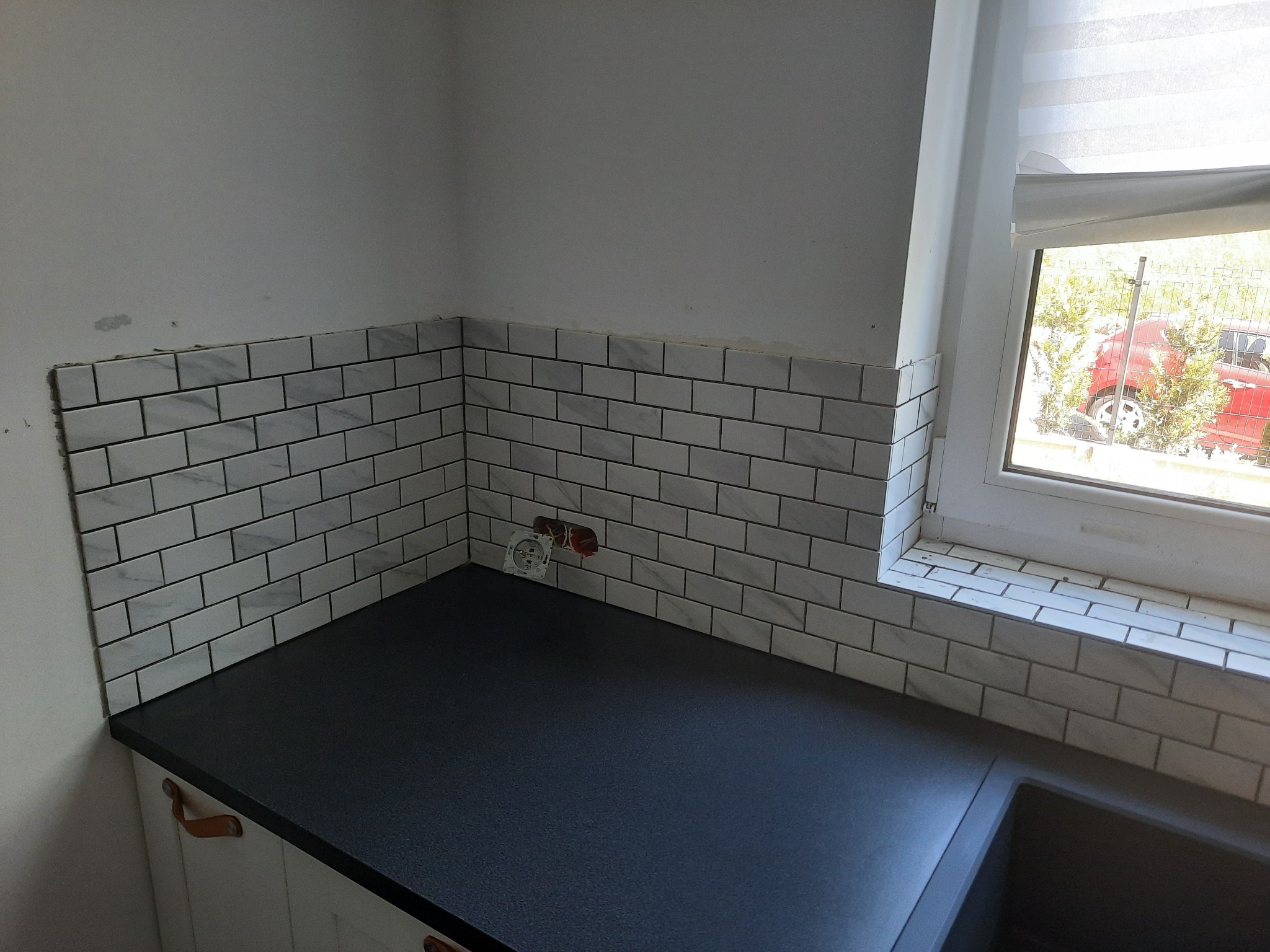 Fragment kuchni w trakcie wykańczania: białe płytki imitujące cegłę na ścianie, czarny blat, widoczne gniazdko elektryczne bez osprzętu, okno z roletą.