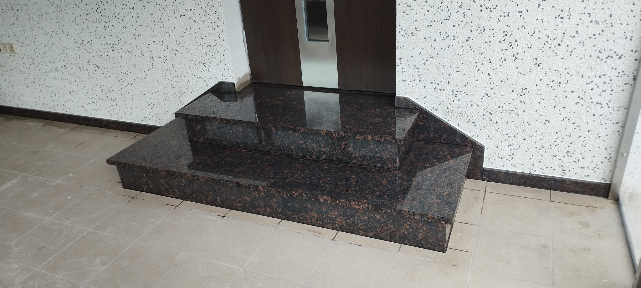 Polerowane, brązowe, dwustopniowe schody wewnętrzne z kamienia, prowadzące do drzwi w Przystajni. Widoczna struktura kamienia i odbicie światła na powierzchni.