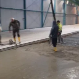 Wylewka betonowa w hali przemysłowej; pracownicy w roboczych ubraniach i żółtych gumowcach wygładzają powierzchnię. Ramię pompy do betonu widoczne w tle.