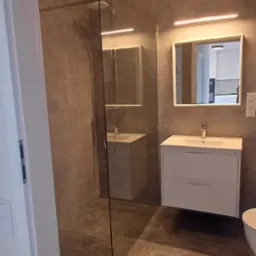 Wykończona łazienka z kabiną prysznicową typu walk-in, umywalką z szafką i lustrem z oświetleniem LED. Płytki w odcieniach szarości na ścianach i podłodze. Widok zza otwartych drzwi.