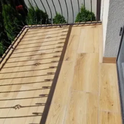 Jasne płytki imitujące drewno na balkonie z czarną, ozdobną balustradą. Widoczne cienie balustrady i fragment zieleni. Wykończony balkon w słoneczny dzień.