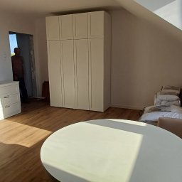 PART TOMASZ BAJDAŁÓW - Jasne wnętrze poddasza ze skosami, z białymi meblami: komoda, szafa. Na podłodze panele. Widoczny fragment sofy i okrągły stół. W tle osoba w drzwiach.