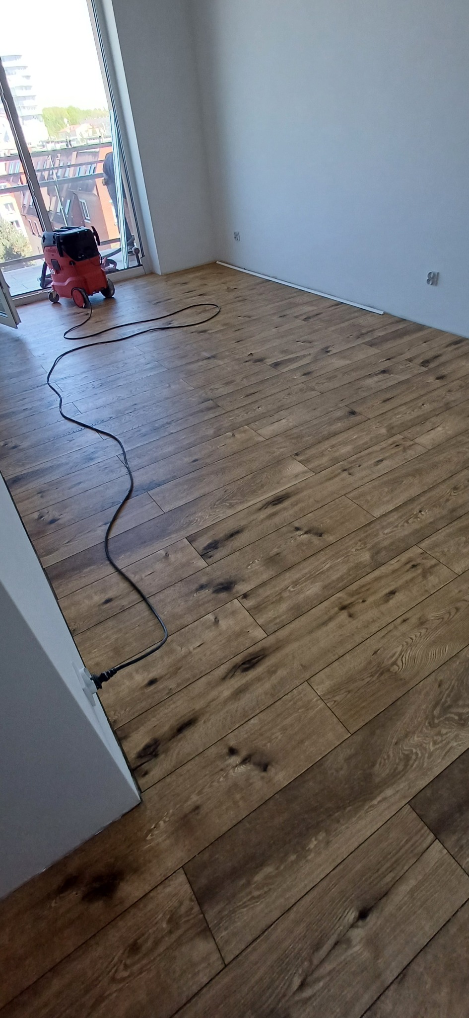 Ukończona podłoga z paneli w nowym mieszkaniu, widoczny odkurzacz przemysłowy i kabel zasilający. Jasne wnętrze z dużym oknem.