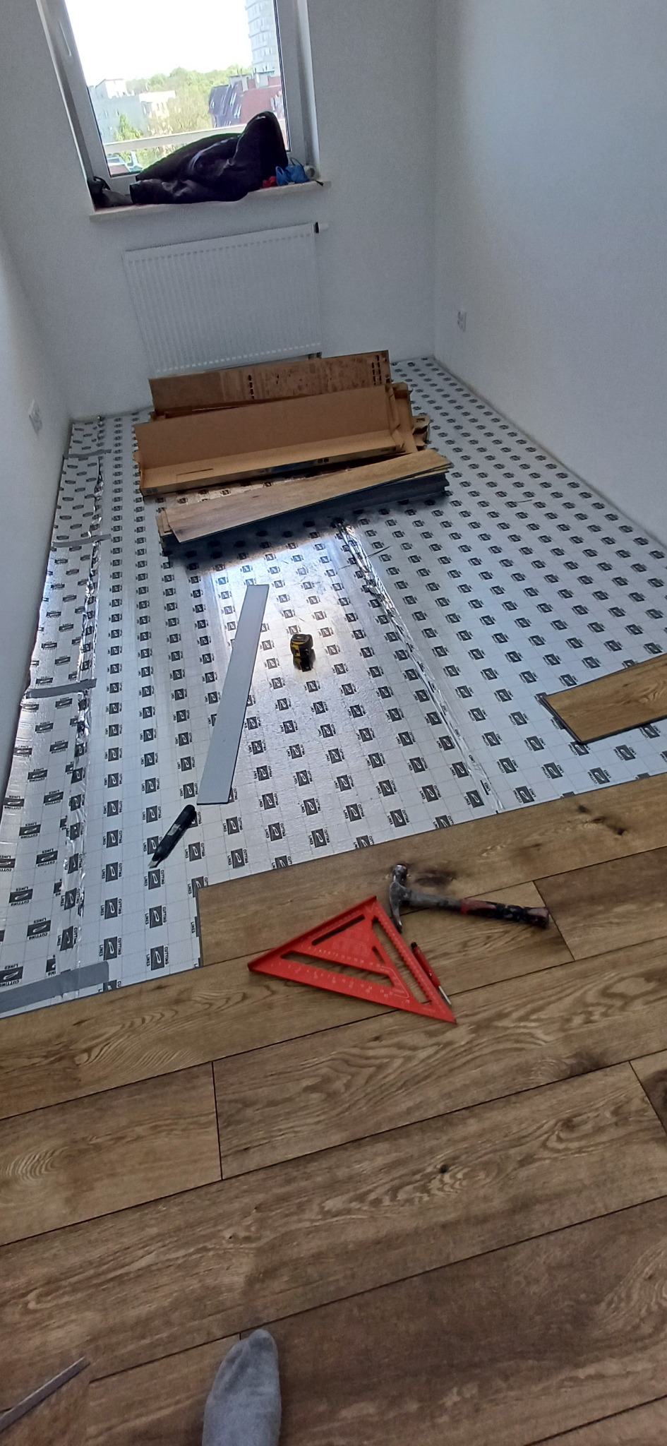 Proces układania paneli podłogowych: widoczny podkład, narzędzia (młotek, kątownik) i częściowo ułożona podłoga z paneli o wzorze drewna. W tle okno.