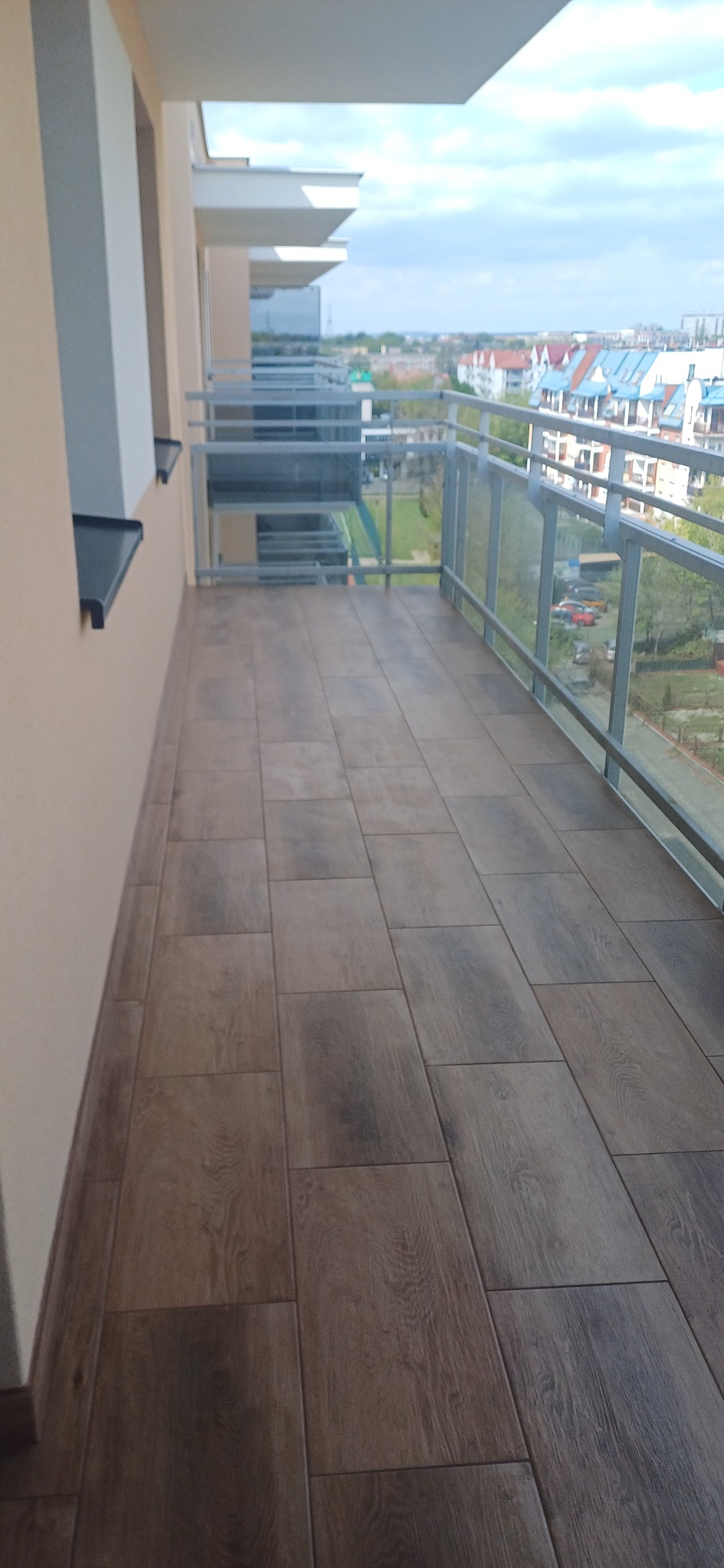 Wykończony balkon z brązowymi płytkami imitującymi drewno, szklane balustrady, widok na miasto w tle. Estetyczne i nowoczesne wykończenie.