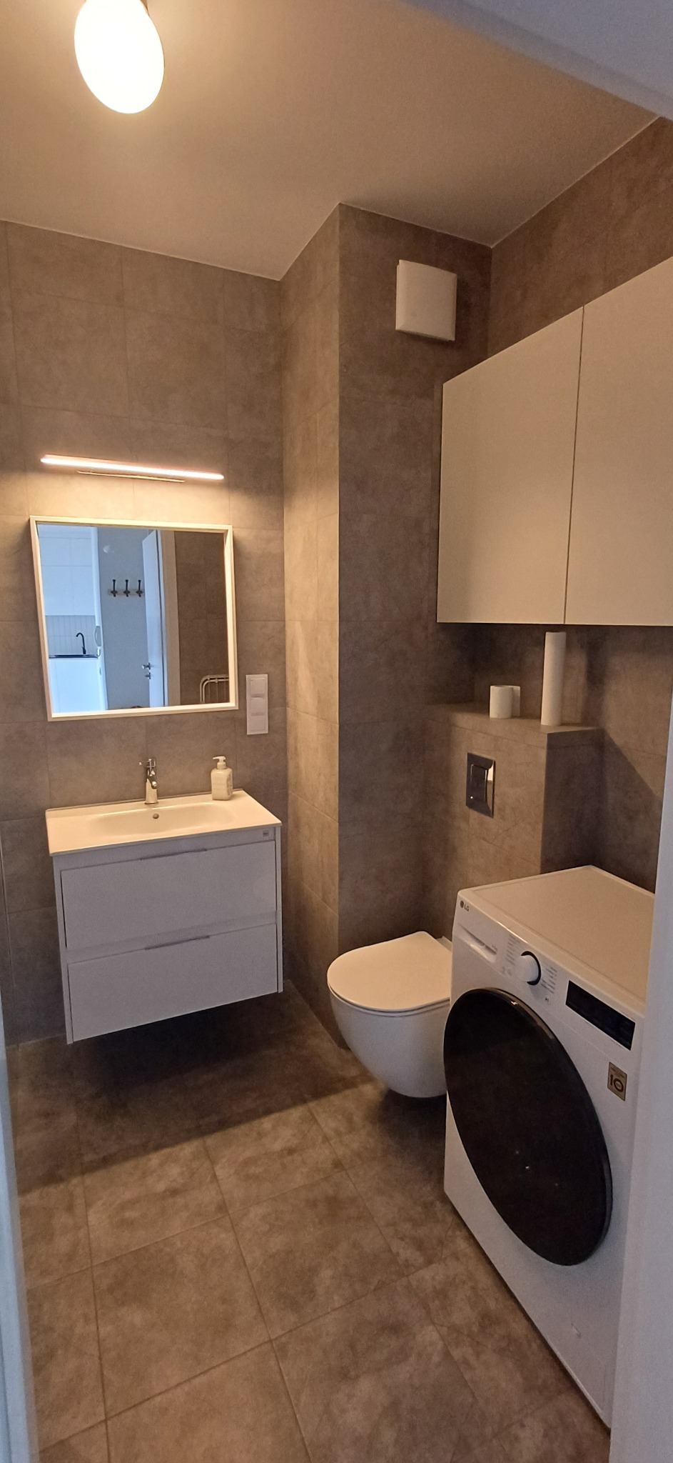 Nowoczesna łazienka z umywalką, lustrem z podświetleniem, toaletą i pralką LG. Stonowana kolorystyka, płytki imitujące kamień. Minimalistyczny design.