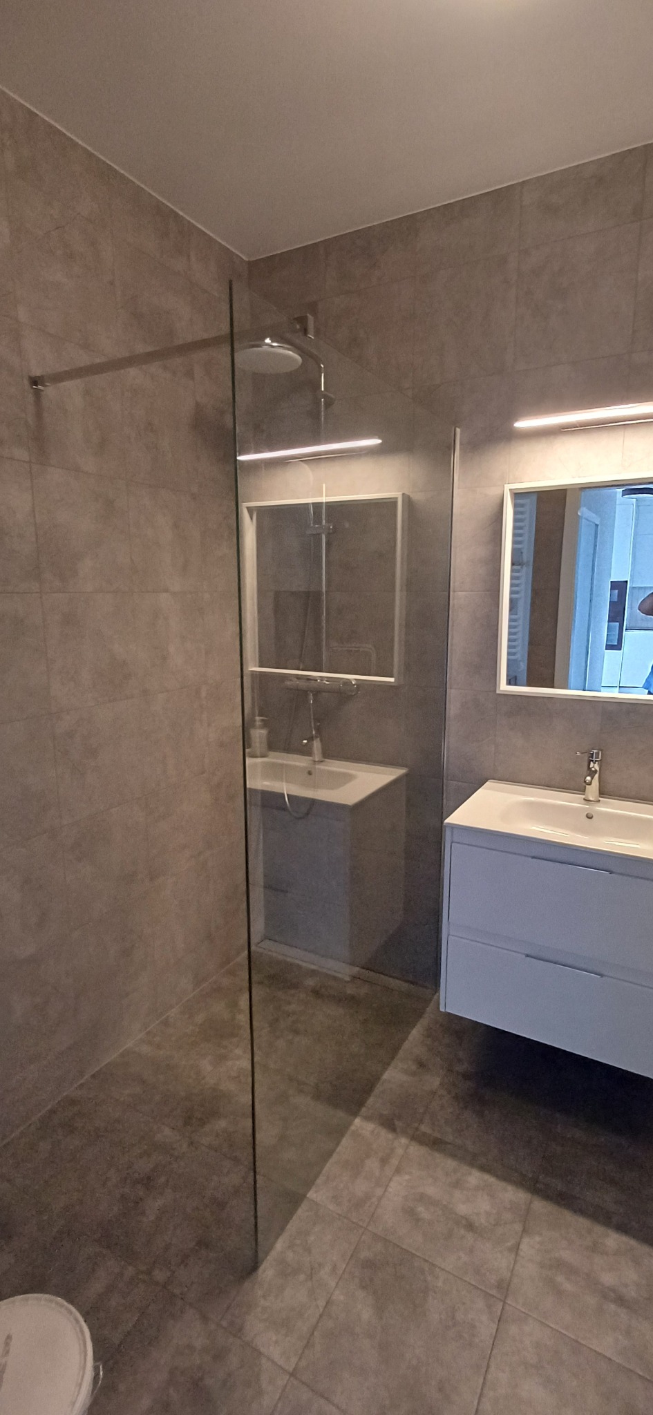 Nowoczesna łazienka z kabiną prysznicową typu walk-in, szarą glazurą i białymi szafkami pod umywalką. Lustro z oświetleniem LED nadaje wnętrzu elegancji.