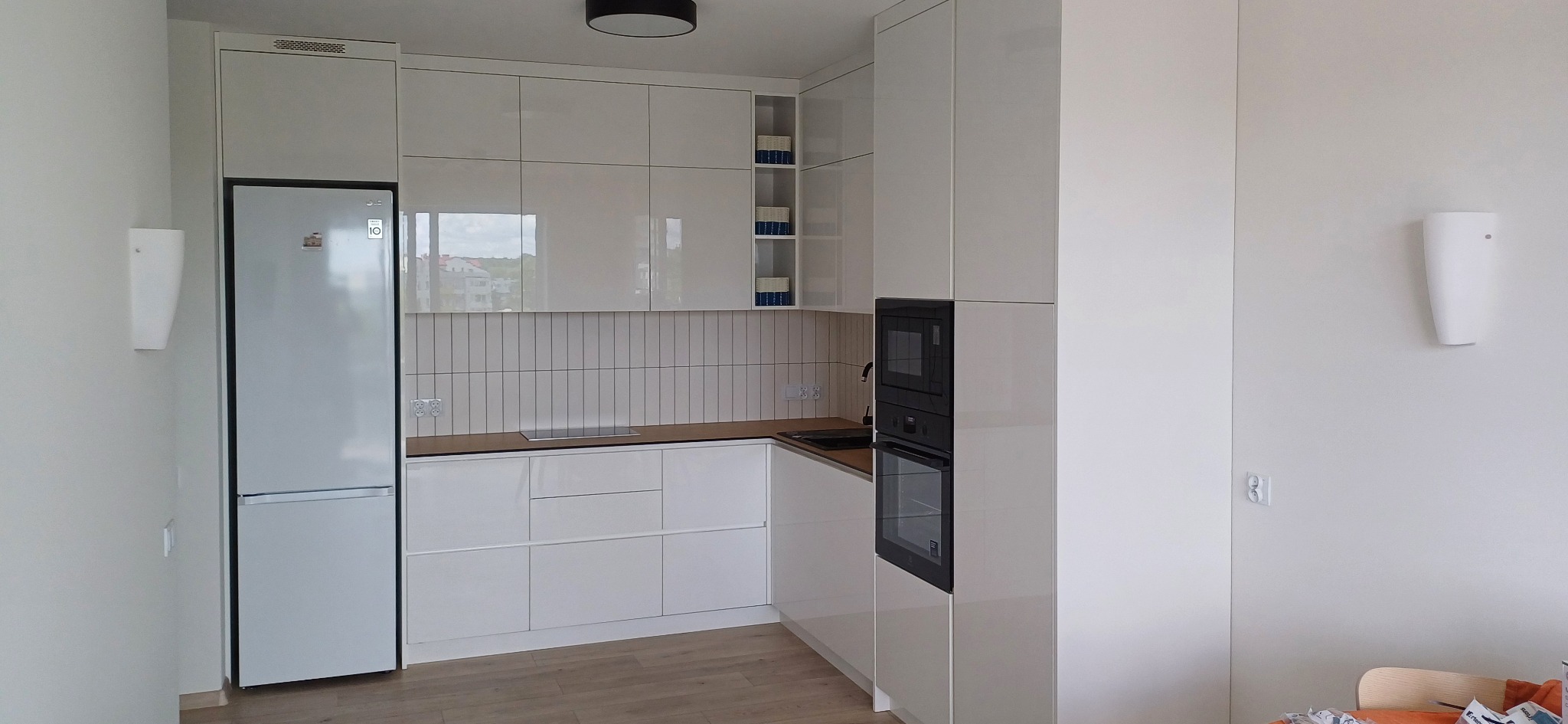 Biała, nowoczesna kuchnia z połyskującymi frontami, pionową glazurą i zabudowaną lodówką. Widoczne gniazdka elektryczne nad blatem i minimalistyczne oświetlenie ścienne.