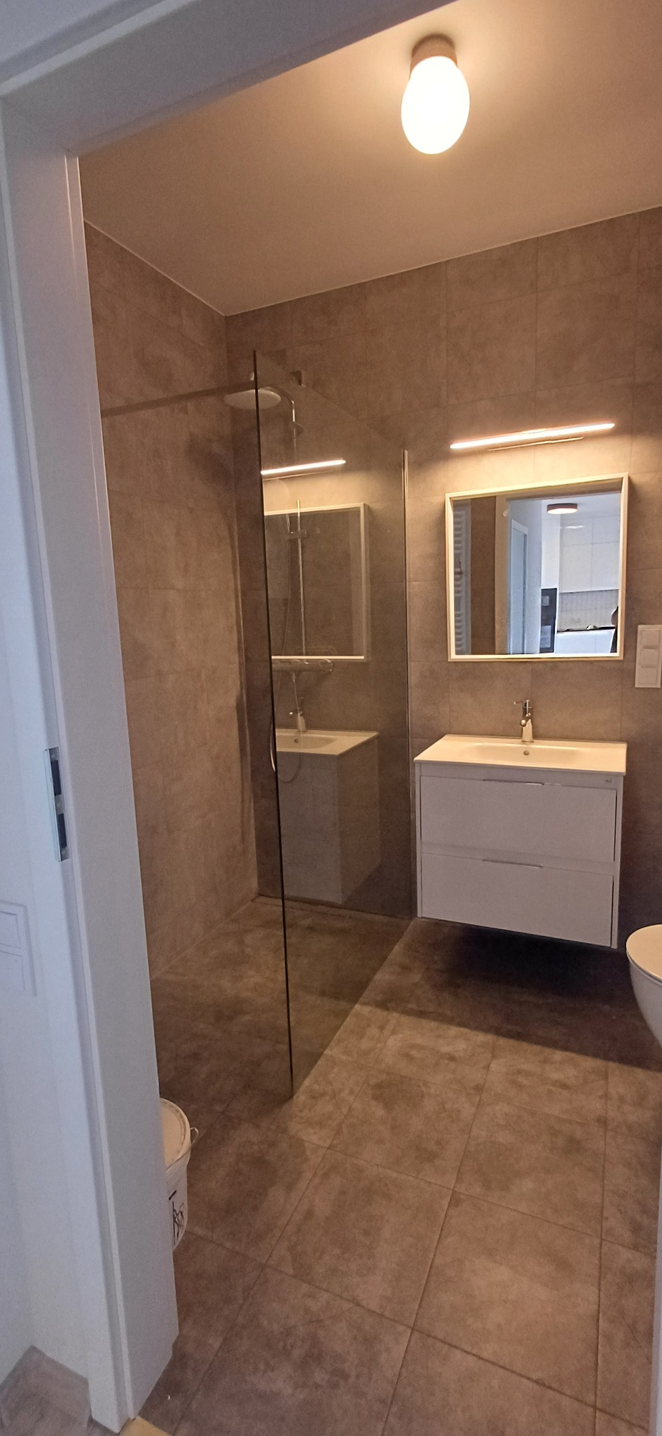 Wykończona łazienka z kabiną prysznicową typu walk-in, umywalką z szafką i lustrem z oświetleniem LED. Płytki w odcieniach szarości na ścianach i podłodze. Widok zza otwartych drzwi.