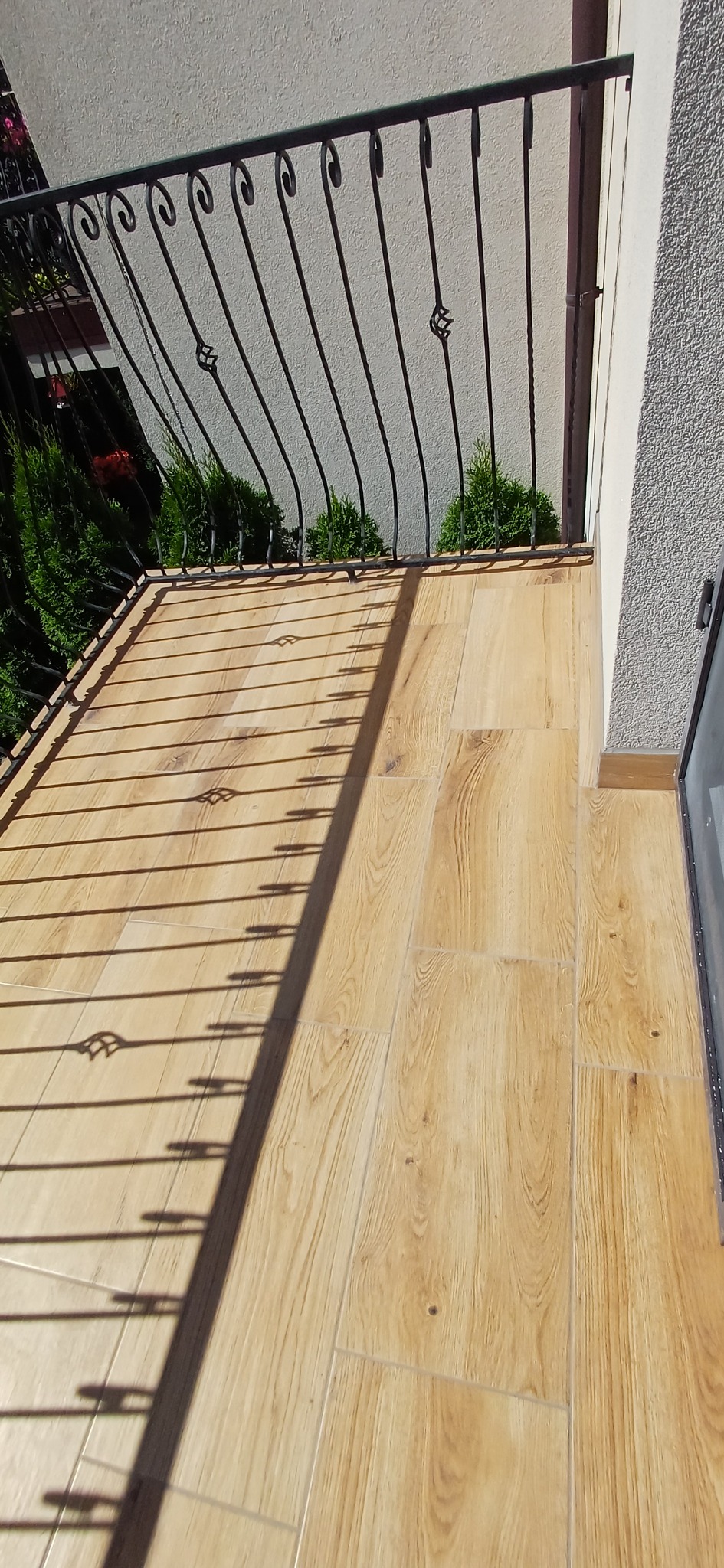 Jasne płytki imitujące drewno na balkonie z czarną, ozdobną balustradą. Widoczne cienie balustrady i fragment zieleni. Wykończony balkon w słoneczny dzień.