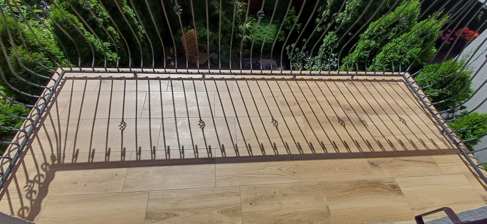 Wykończony balkon z drewnopodobną terakotą i metalową balustradą, z widokiem na zieleń. Cienie balustrady tworzą wzór na podłodze.
