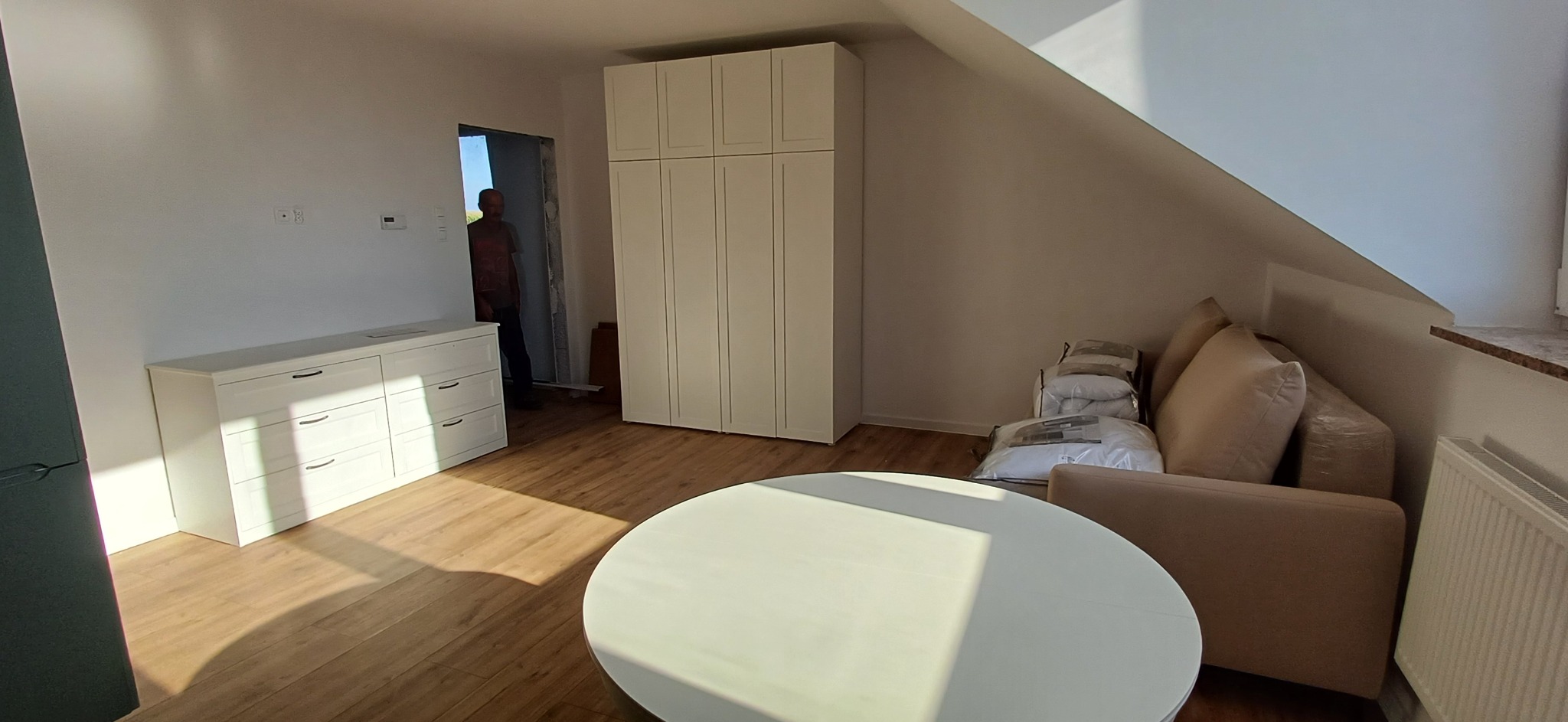 Jasne wnętrze poddasza ze skosami, z białymi meblami: komoda, szafa. Na podłodze panele. Widoczny fragment sofy i okrągły stół. W tle osoba w drzwiach.