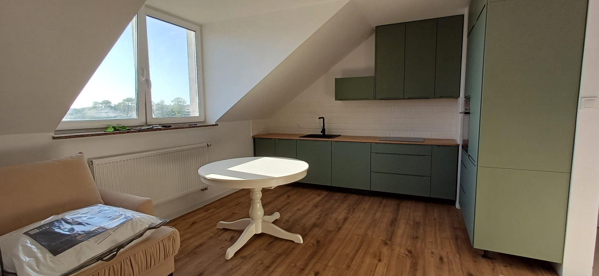 Kuchnia na poddaszu z zielonymi szafkami i białymi płytkami, okrągły stół w centrum, okno dachowe z widokiem na zieleń, drewniana podłoga, fotel z zapakowaną pościelą.
