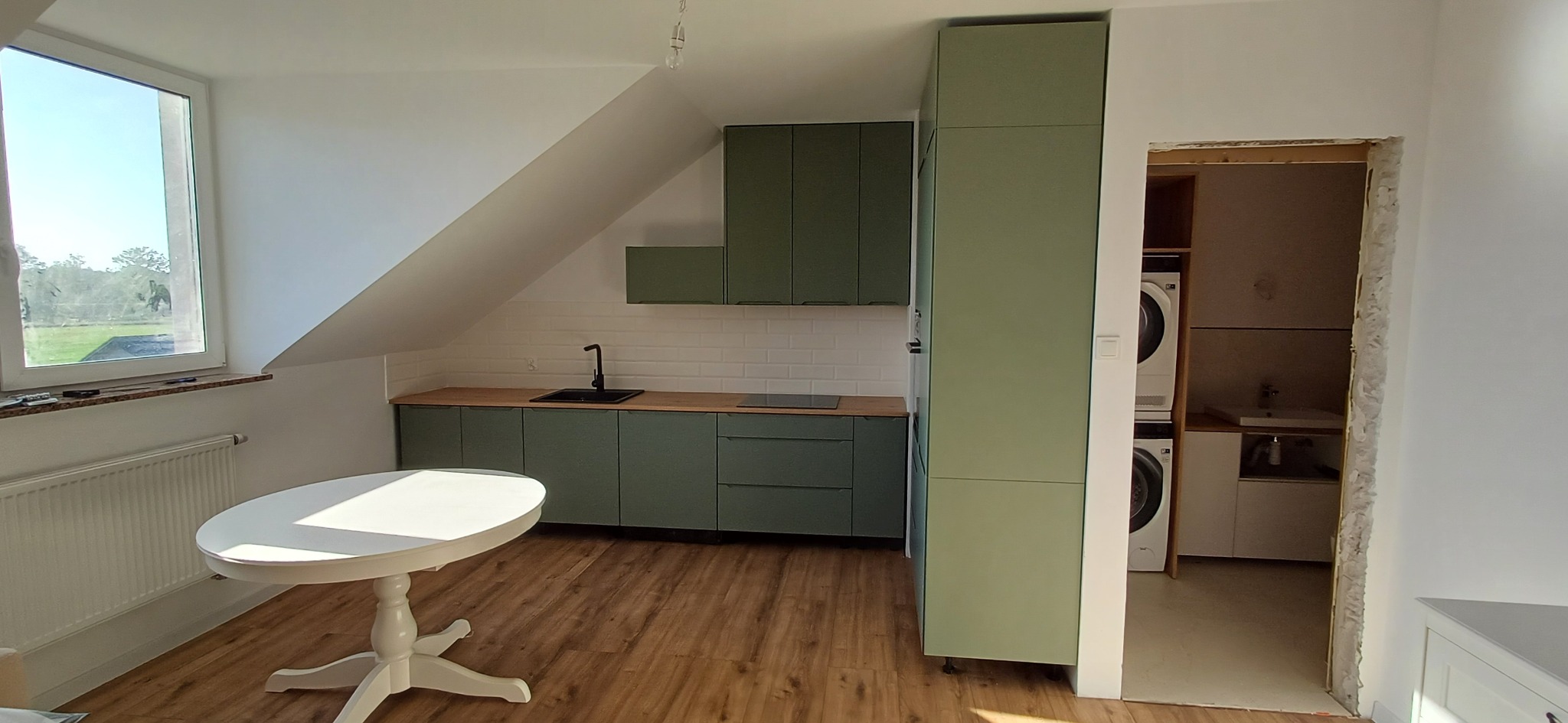 Kuchnia na poddaszu z zielonymi szafkami, drewnianym blatem i białymi płytkami. Okrągły stół w centrum, pralka w tle. Naturalne światło z okna.