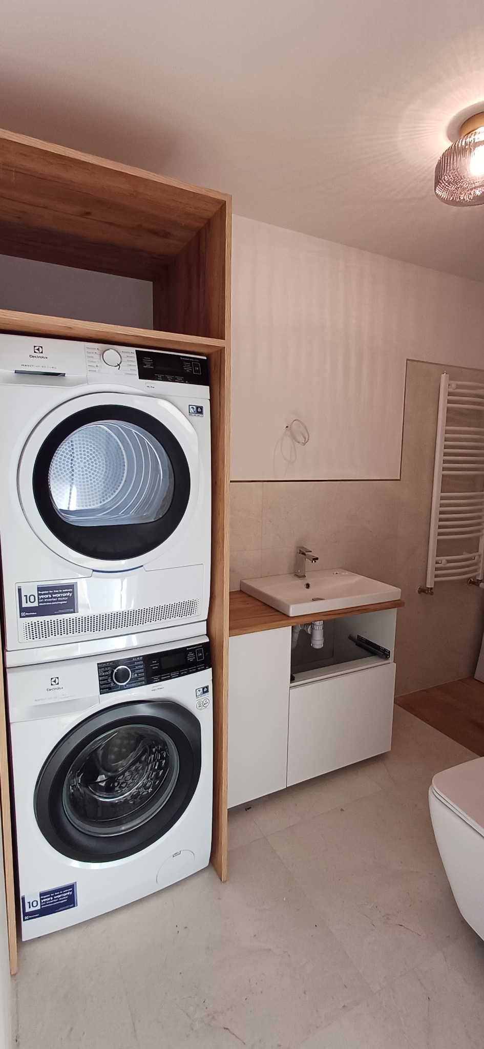 Nowoczesna łazienka z pralką i suszarką Electrolux w zabudowie drewnianej, umywalką z szafką i grzejnikiem drabinkowym w tle.