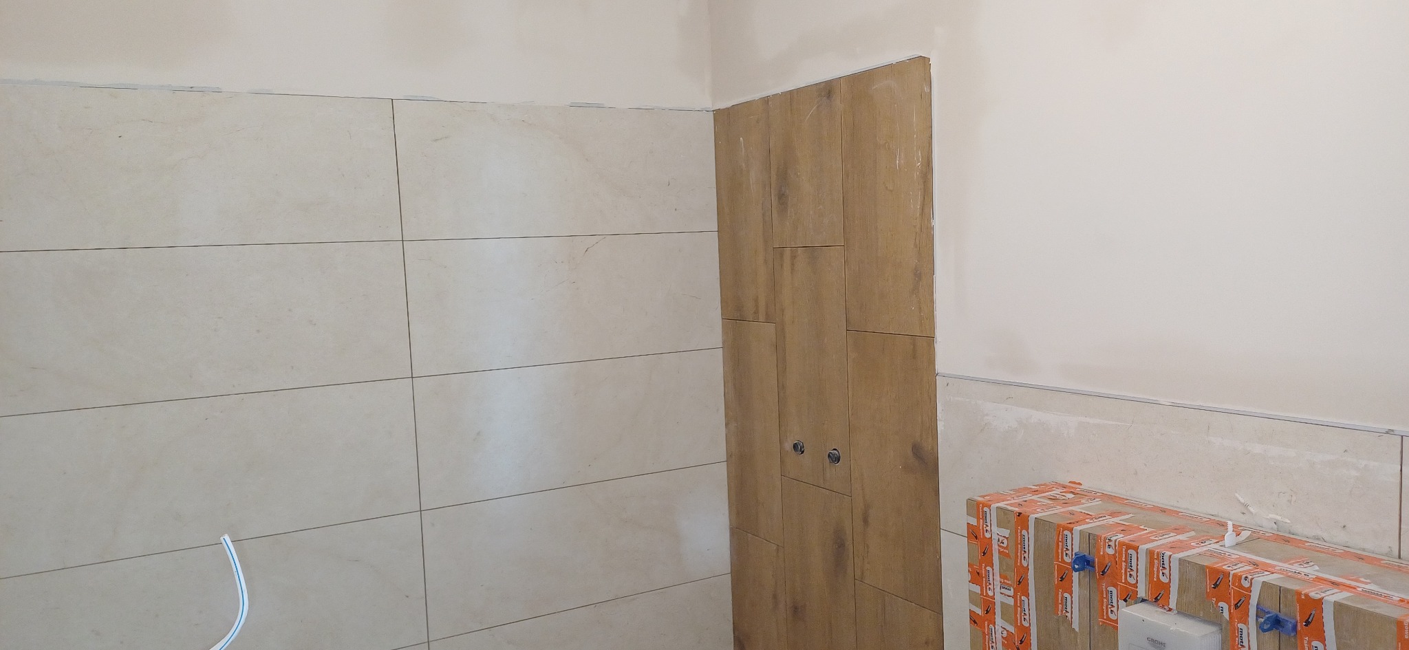 Wykończona ściana z beżowych płytek imitujących kamień, obok fragment ściany wyłożonej drewnopodobnymi panelami z dwoma chromowanymi punktami mocowania.