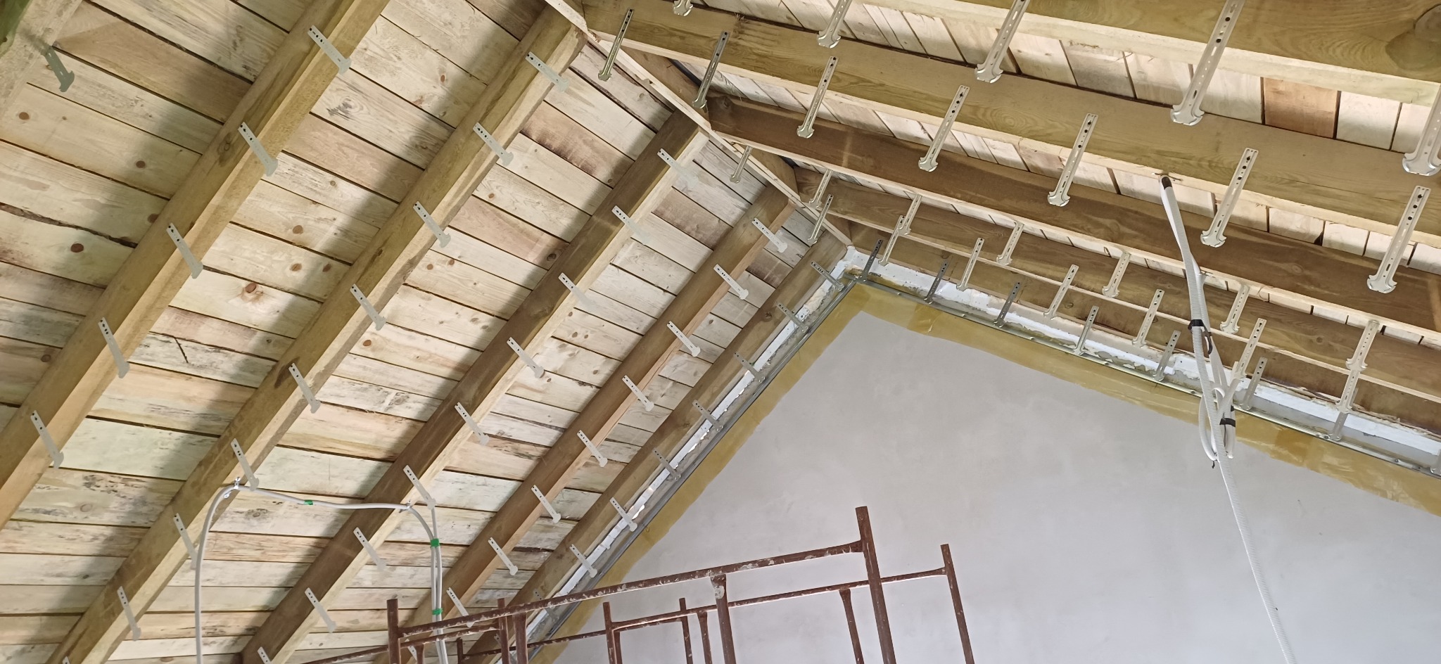 Drewniany szkielet poddasza z zamocowanymi metalowymi uchwytami, przygotowany do ocieplenia. Widoczne przewody instalacyjne i fragment rusztowania.