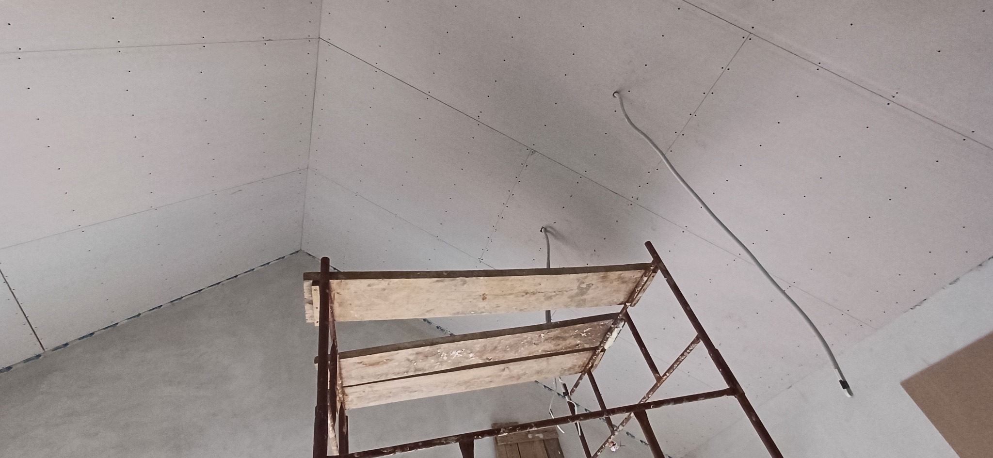 Poddasze z zamontowaną płytą gipsowo-kartonową, widoczne ślady po wkrętach i rusztowanie. Kabel zwisa z sufitu. Ujęcie z dołu, podkreślające skos dachu.