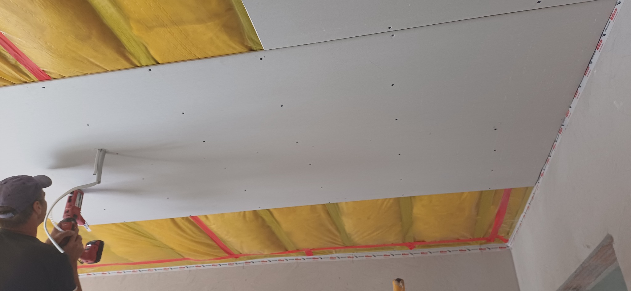 Montaż płyty gipsowo-kartonowej na suficie z widoczną izolacją. Pracownik w trakcie pracy przy użyciu wkrętarki, widoczny fragment pomieszczenia i oświetlenie.