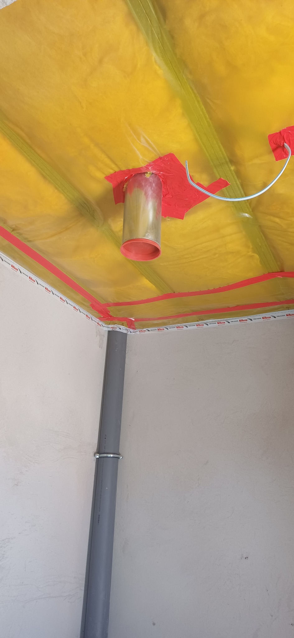 Żółta folia paroizolacyjna na suficie z rurą wentylacyjną i taśmą uszczelniającą, szara rura kanalizacyjna przy ścianie. Detal instalacji w trakcie prac wykończeniowych.