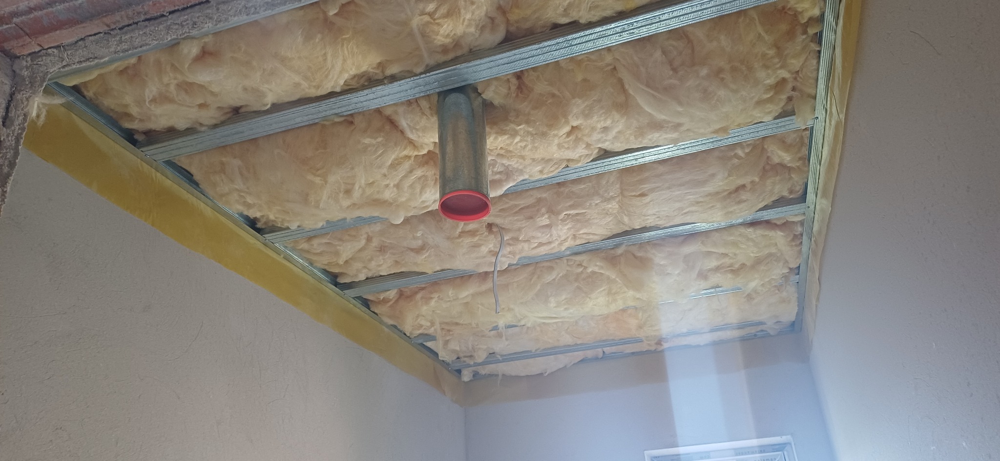 Ocieplone poddasze wełną mineralną z widoczną konstrukcją metalową i rurą wentylacyjną. Żółta taśma uszczelniająca na obrzeżach.