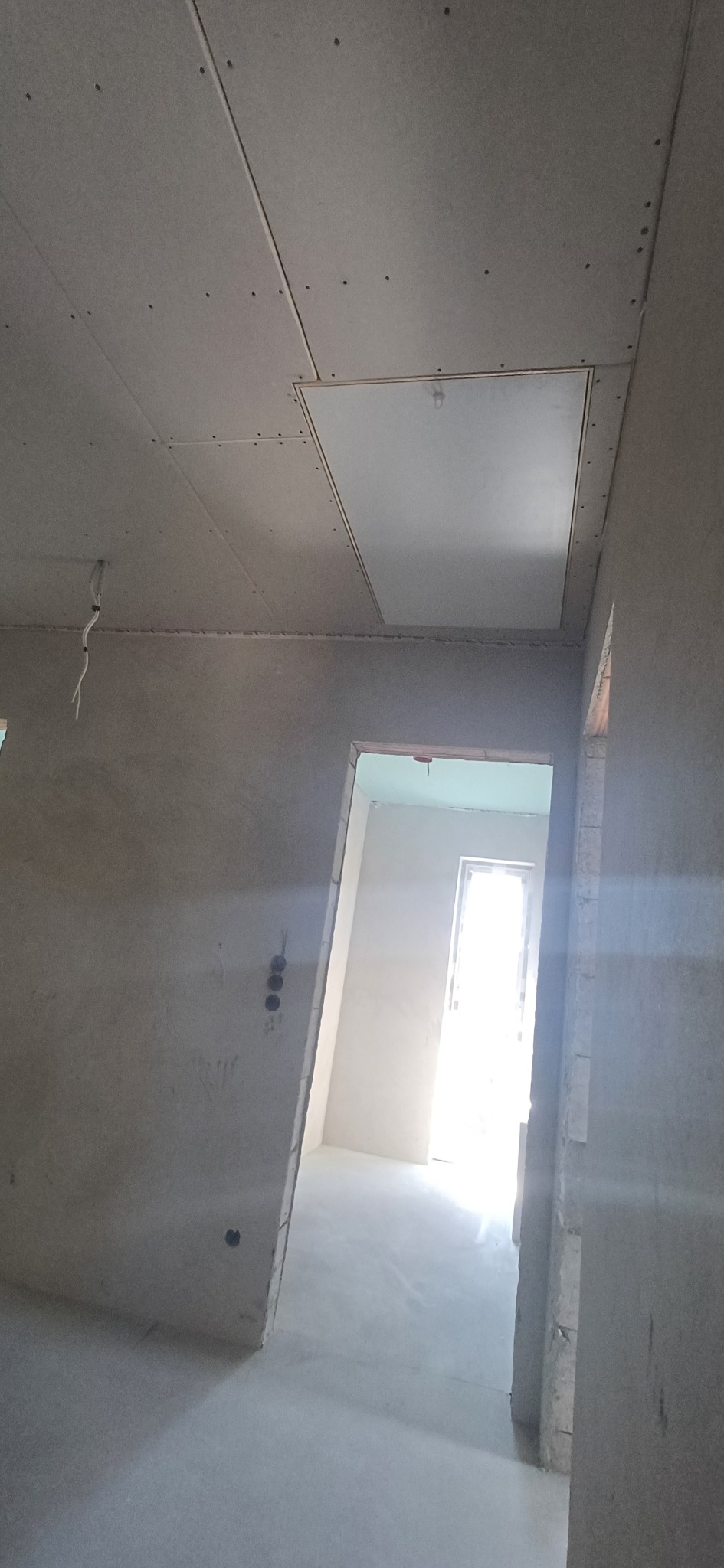 Montaż płyty karton gips na suficie z widocznym otworem rewizyjnym. Ściany w trakcie wykańczania, widoczne puszki elektryczne. W tle widok na kolejne pomieszczenie.