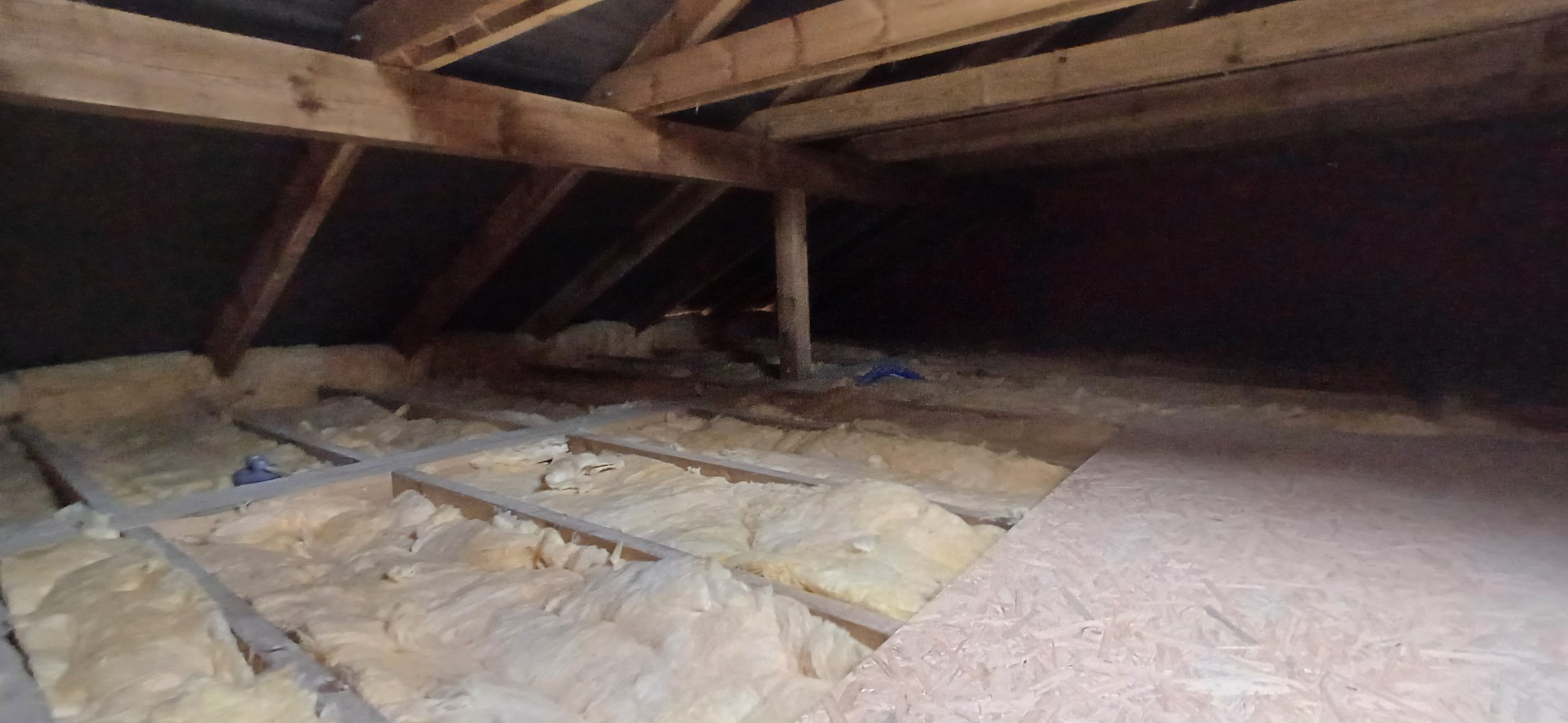 Poddasze z widoczną izolacją z wełny mineralnej między drewnianymi belkami, częściowo zakryte płytą. Widok z góry na ocieplony strych.