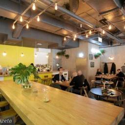 ŚWIDERSKI PRACOWNIA PROJEKTOWA Świderski Sławomir - Wnętrze budynku usługowego: Długa, jasna, drewniana lada barowa z wysokimi krzesłami, za nią bar z butelkami i roślinami, w tle siedzący goście przy stolikach, surowy sufit z widocznymi...