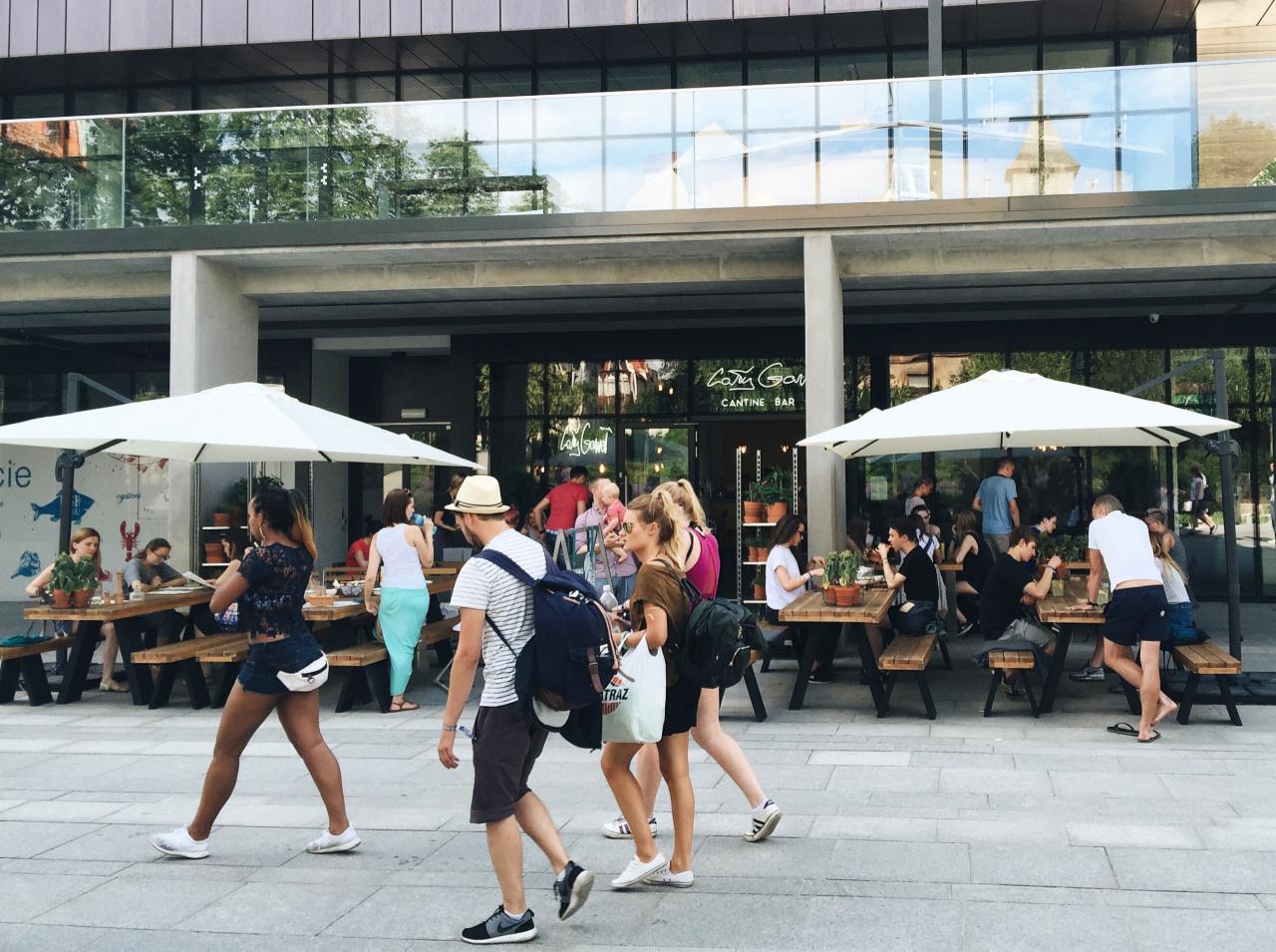 Nowoczesny budynek usługowy z przeszkloną fasadą, przed którym znajdują się drewniane stoliki z parasolami i siedzący goście.