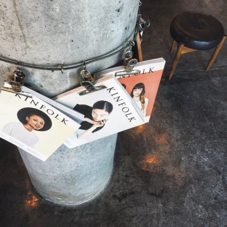 Metalowy kubeł z przypiętymi okładkami magazynu Kinfolk, obok czarny stołek na trzech nogach, surowa podłoga