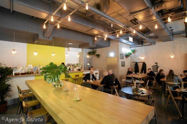 Wnętrze budynku usługowego: Długa, jasna, drewniana lada barowa z wysokimi krzesłami, za nią bar z butelkami i roślinami, w tle siedzący goście przy stolikach, surowy sufit z widocznymi...