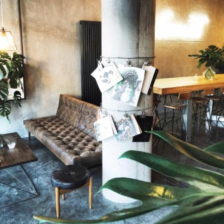Wnętrze kawiarni w stylu industrialnym z betonową kolumną ozdobioną grafikami, skórzaną sofą z pikowaniem, drewnianym stołem i roślinami doniczkowymi.