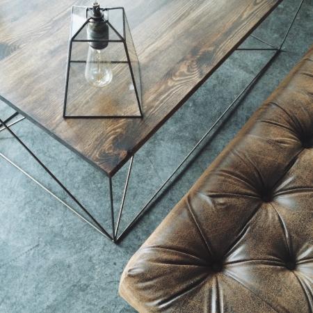 Stylowy stolik kawowy z ciemnego drewna z metalową podstawą i dekoracyjną lampą w kształcie klatki, obok skórzany puf z guzikami, widok z góry.