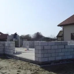 Wznoszenie ścian parteru z bloczków na fundamencie domu jednorodzinnego, widoczni pracownicy w kombinezonach, w tle budynek z czerwoną dachówką.
